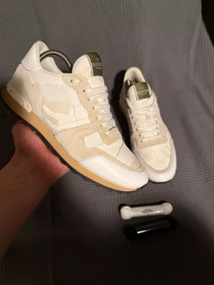Valentino rockrunner  - Snygga sneakers från Valentino i beige och vitt med camomönster. Skorna har detaljer i mocka och läder, samt en olivgrön logga på plösen. Yttersulan är i gummi med beige och svart färg. Perfekta för dig som gillar exklusiva och trendiga sneakers.