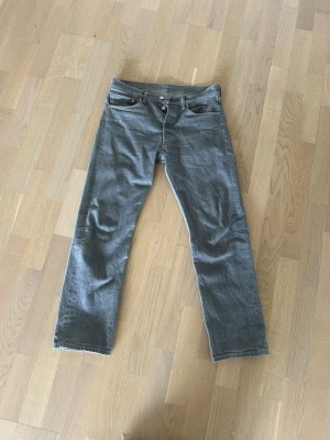 Raka Levis 501 - Säljer ett par grå jeans från Levi's med klassisk femficksmodell och raka ben. Jeansen har knappgylf och detaljer i metall. Perfekta för en avslappnad och stilren look.