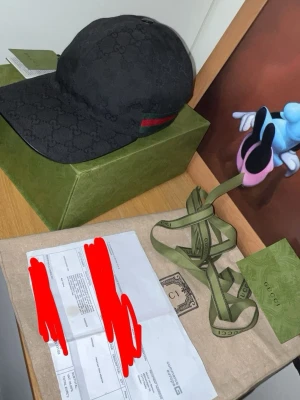 Svart Gucci keps med GG-mönster - Snygg svart keps från Gucci med klassiskt GG-monogram över hela kepsen och en grön-röd rand på sidan. Kepsen är tillverkad i tyg och har en böjd skärm. Perfekt accessoar för dig som vill ha en exklusiv och trendig look.