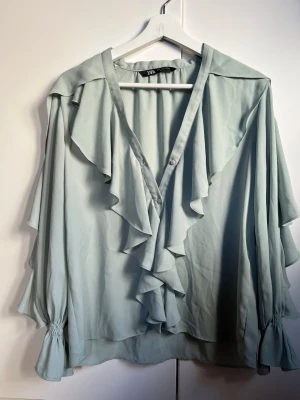 Ljusblå volangblus från Zara - Snygg ljusblå blus från Zara med volangdetaljer längs urringningen och framtill. Blusen har långa, vida ärmar med resår vid ärmsluten och är tillverkad i ett tunt, mjukt tyg som ger ett flowy intryck. Perfekt för dig som gillar romantiska detaljer.