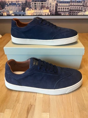 Mörkblå sneakers från Axel Arigato - Stilrena mörkblå sneakers i mocka från Axel Arigato med vit sula och guldfärgad logga på hälen. Skorna har låga skaft och bruna detaljer på insidan. Perfekta för dig som gillar minimalistisk design och premiumkänsla.