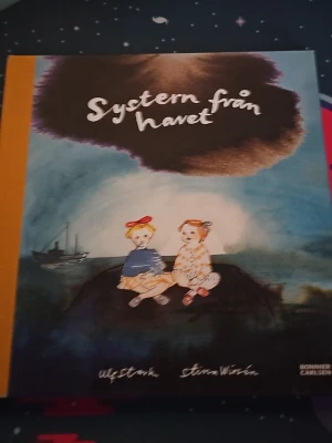 Systern från havet bok - En illustrerad bok med titeln 'Systern från havet' av Ulf Stark och Stina Wirsén. Omslaget har en blå och brun akvarellbakgrund med två tecknade flickor som sitter tillsammans vid havet. Boken har en gul rygg och mjuka pärmar.