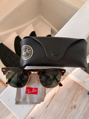 Ray-Ban Clubmaster solglasögon brun - Snygga Ray-Ban Clubmaster solglasögon med brunsköldpaddsmönstrad båge och guldfärgade detaljer. Klassisk retroform med mörka glas. Kommer med originalfodral i svart skinn och putsduk med Ray-Ban-logga.