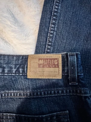Blå bootcut jeans från Mustang - Säljer ett par klassiska blå bootcut jeans från Mustang med snyggt läderpatch bak. Jeansen har fem fickor, normal midja och är tillverkade i slitstarkt denim. Perfekt för dig som gillar tidlös stil och bekväma jeans.