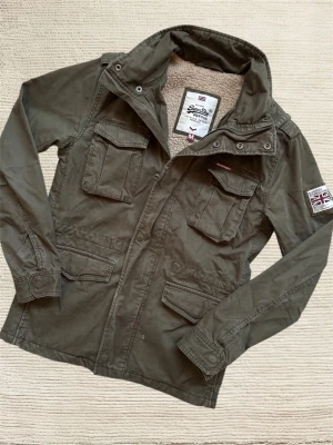 SUPERDRY FIELDJACKET - En mycket trevlig fieldjacket av märket Superdry i en extremt snygg grön färg! Pytteliten defekt på sista bilden. Hör av dig vid frågor!