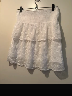 Vit volangkjol med broderade blommor - Söt vit kjol med två volanger och broderade blommor. Kjolen har en smockad midja som ger en bekväm passform och ett luftigt, romantiskt fall. Perfekt till sommaren och lätt att matcha med olika toppar.