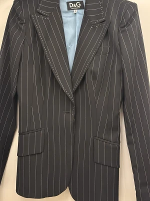 Svart D&G kavaj med kritstreck - Snygg svart kavaj från Dolce & Gabbana med vita kritstrecksränder och ljusblått foder. Klassisk passform med spetsiga slag, två fickor med lock och en bröstficka. Perfekt för en stilren och trendig look.