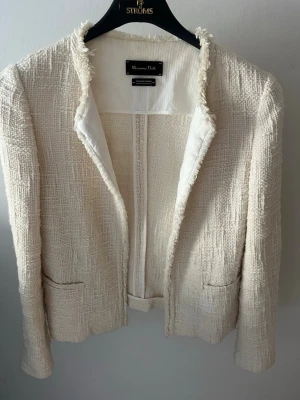 Krämvit bouclé kavaj Massimo Dutti - Stilren krämvit bouclé kavaj från Massimo Dutti med råa kanter och två fickor framtill. Kavajen har en öppen design utan knappar och är helfodrad i vitt tyg. Materialet är strukturerat och ger en exklusiv känsla.