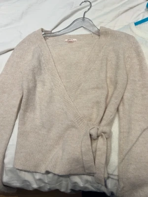 Beige omlottkofta med knytband - Mysig beige omlottkofta med knytband i midjan och ribbade muddar. Koftan har en djup V-ringning och långa ärmar, perfekt för lager på lager. Materialet ser ut att vara mjukt och lite fluffigt, vilket ger en avslappnad och trendig look.