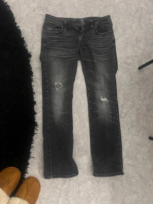 Svarta slitna jeans med raka ben - Säljer ett par svarta jeans med slitningar på båda benen och snyggt tvättad look. Jeansen har raka ben och klassisk femficksmodell. Perfekta för dig som gillar en avslappnad och trendig stil.