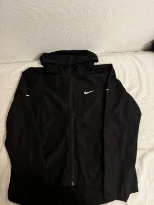 Svart Nike vindjacka med huva - Snygg svart vindjacka från Nike med hel dragkedja och huva. Jackan har reflekterande detaljer på ärmarna och den klassiska Nike-loggan på bröstet. Tillverkad i lätt och vindtätt material, perfekt för blåsiga dagar.