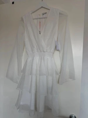 Vit omlottklänning med volanger - Supersöt vit kortklänning från bubbleroom med omlott framtill, långa transparenta ärmar och flera lager volanger i kjolen. Smockad midja ger en snygg passform och klänningen har en lätt och luftig känsla, perfekt för varma dagar, som studentklänning eller till skolavslutning 