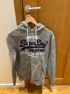 Grå hoodie från Superdry med tryck - Snygg grå hoodie från Superdry med stort mörkblått och vitt tryck på bröstet. Klassisk känguruficka framtill, vita dragsnören i huvan och mjukt material på insidan. Perfekt för en avslappnad och trendig stil.