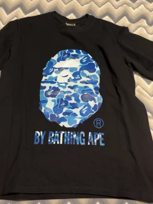 Svart BAPE t-shirt med blått camotryck - Säljer en svart t-shirt från BAPE med det ikoniska apansiktet i blått camomönster på bröstet och texten 'BY BATHING APE' under. T-shirten har rund hals och korta ärmar. Perfekt för dig som gillar streetwear och vill sticka ut.