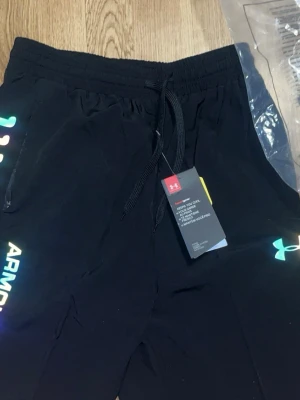 Svarta shorts från Under Armour - Svarta träningsshorts från Under Armour med färgglada detaljer och logga på benet. Shortsen har elastisk midja med snörning och är tillverkade i ett lätt och funktionellt material som passar perfekt för träning.
