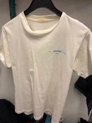 Vit Off-White t-shirt med tryck - Säljer en vit t-shirt från Off-White med blå text på bröstet och stort grafiskt tryck i blått, rosa och brunt på ryggen. Klassisk passform och rund hals. Perfekt för dig som gillar streetwear och vill sticka ut med en ikonisk design.