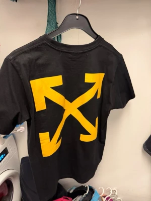 Svart Off-White t-shirt med tryck - Svart t-shirt från Off-White med ikoniska gula pilar på ryggen och konstmotiv med gula detaljer framtill. Klassisk rund hals och korta ärmar. Tillverkad i mjuk bomull för en skön känsla och streetwear-vibe.