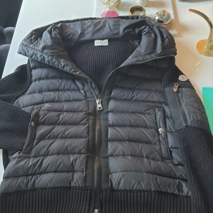 Svart cardiganjacka från Moncler - Snygg svart cardiganjacka från Moncler med quiltade partier och stickade ärmar. Jackan har dragkedja framtill, fickor med knappstängning och en huva. Perfekt för dig som vill ha en sportig och trendig look med exklusiv känsla. Perfekt för kommande vår! Priset är inte hugget i sten!! kvitto finns att få bild på 