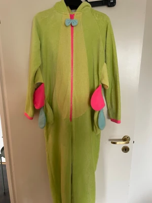 Grön larv onesie med färgglada detaljer - Rolig och mysig larvdräkt i onesie-modell, perfekt för maskerad eller cosplay. Dräkten är grön med dragkedja framtill, har huva med larvansikte och antenner samt färgglada detaljer i rosa, blått, gult, orange och lila. Tillverkad i mjukt fleece-liknande material.