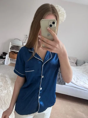 Mörkblå satinpyjamas med vita detaljer - Snygg mörkblå pyjamaströja i satin med korta ärmar och vita kontrastsömmar. Klassisk krage, bröstficka och knäppning framtill med vita knappar. Jag köpte tröjan begagnat men jag har själv nästan aldrig använt den 💕