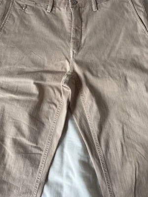 Beige chinos från Replay - Snygga beige chinos från Replay med klassisk rak passform och diskreta fickor fram. Tillverkade i ett mjukt bomullsmaterial som ger en skön känsla. Perfekta för dig som vill ha en stilren och enkel look.