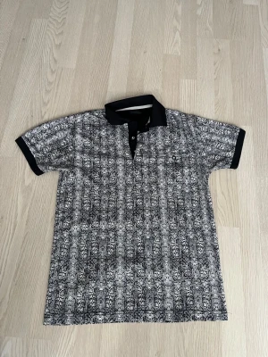 mönstrad pikéskjorta - Säljer en svartvit pikéskjorta med korta ärmar och ett detaljerat mönster över hela plagget. Skjortan har svart krage och svarta muddar vid ärmsluten samt knappar framtill. Perfekt för dig som gillar unika prints och stilren design.