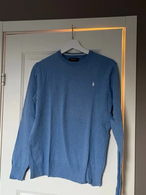 Blå stickad tröja Polo Ralph Lauren - Snygg blå stickad tröja från Polo Ralph Lauren i mjuk pima bomull. Klassisk rund halsringning, ribbade muddar och den ikoniska vita logon broderad på bröstet. Perfekt till jeans eller chinos för en clean look.