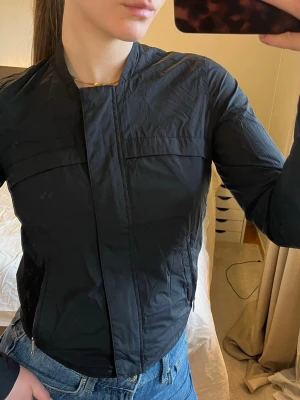 Svart vind / träningsjacka - storlek xs - Svart vårjacka med ”sportigt” utseende med två sidofickor och bröstfickor, öppningsbara ärmslut - Peak Performance   Storlek xs