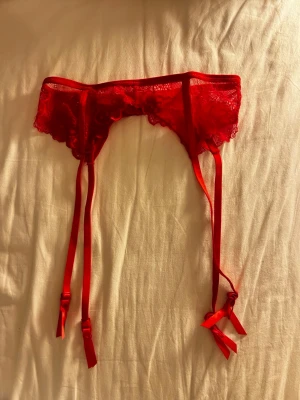 Röd spetsigt strumpebandshållare - Snygg röd strumpebandshållare i spets med fyra justerbara band och dekorativa rosetter. Tillverkad i ett tunt, genomskinligt material med fina spetsdetaljer för en extra feminin look. Aldrig använd. Från Hunkemöller.