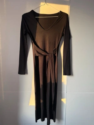 Svart ribbad långklänning med knyt - Svart långklänning i ribbad bomull med v-ringning och långa ärmar. Klänningen har ett knytband i midjan som ger en snygg siluett. Perfekt för dig som gillar stilrena och bekväma plagg.