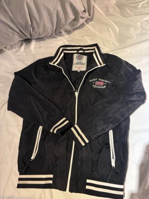 Svart vindjacka Saint Vincent Polo Club - Snygg svart vindjacka från Saint Vincent Polo Club med vita detaljer och dragkedjor. Jackan har broderad logga på bröstet, ståkrage med text och randiga muddar vid ärmslut och nederkant. Perfekt för en sportig och stilren look. Storlek 150