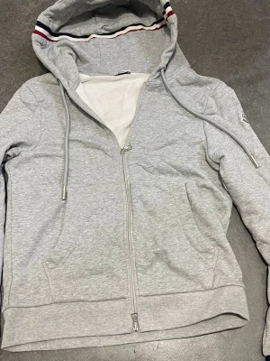 Grå zip-hoodie från Moncler - Snygg grå hoodie med dragkedja från Moncler. Huva med dragsnören och coola röd-blå-vita detaljer på insidan av huvan och ärmslut. Mjukt material och två fickor framtill. Perfekt för en avslappnad och trendig look.
