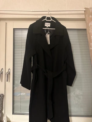 Svart kappa från NA-KD med skärp - Stilren svart kappa från NA-KD med klassisk krage och knytband i midjan. Lång modell med raka ärmar och mjukt tyg som ger en snygg siluett. Perfekt för dig som vill ha en enkel men trendig look.