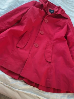 Röd trenchcoat med huva från Lindex - Stilren röd trenchcoat från Lindex med huva, stora knappar framtill och två fickor. Jackan har en klockad form och dekorativa detaljer i ryggen med knappar och veck. Perfekt för dig som vill sticka ut i höst och vår.