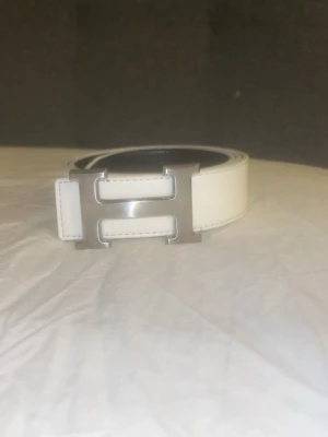 Bälte H spänne vändbart - Snyggt vändbart bälte i vitt och svart med ett stort H-format metallspänne. Perfekt accessoar för att lyfta din outfit och ge en clean look. Bältet har tydliga sömmar och är gjort i skinn.