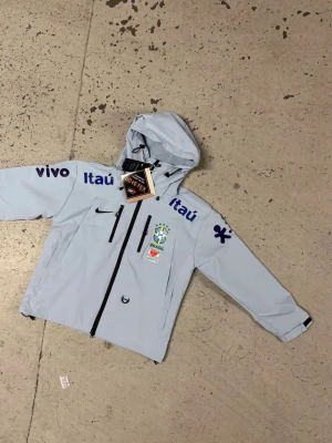 Nike Brasil vindjacka ljusblå - Ljusblå vindjacka från Nike med Brasil-logga och flera sponsortryck i blått. Jackan har huva med dragsko, svarta dragkedjor framtill och på bröstet samt justerbara ärmslut. Tillverkad i ett lätt och vattenavvisande material, perfekt för blåsiga dagar.