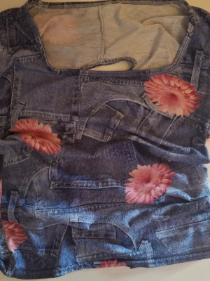Blommig topp med jeansmönster och cut-outs - Trendig topp med blått jeansmönster och stora rosa blommor. Toppen har flera cut-outs på ryggen som ger en edgy look. Materialet känns mjukt och stretchigt, perfekt för dig som vill sticka ut. Rund halsringning och korta ärmar.
