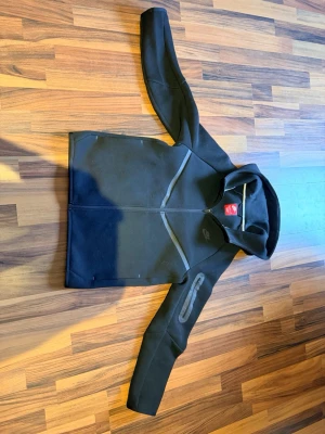 Svart hoodie från Nike med dragkedja - Säljer en svart hoodie från Nike med hel dragkedja framtill och huva. Hoodien har grå detaljer längs bröstet och en praktisk ficka med dragkedja på ena ärmen. Perfekt för dig som gillar sportig och clean stil. Kan gå ner i pris vid snabb affär