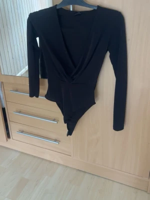 Svart långärmad body med djup urringning - Snygg svart body med lång ärm och djup omlott-urringning framtill. Tight passform som framhäver figuren och passar perfekt till både jeans och kjol. Tillverkad i stretchigt material för skön känsla hela dagen.