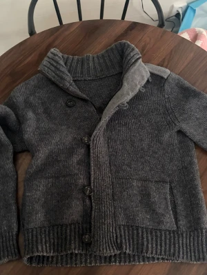 Mörkgrå stickad kofta med krage - Mysig mörkgrå stickad kofta med bred ribbad krage och knäppning framtill. Koftan har två fickor, ribbade muddar och axeldetaljer med knappar. Perfekt för lager på lager och chill stil.