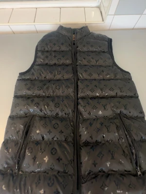 Svart dunväst från Louis Vuitton - Snygg svart dunväst från Louis Vuitton med ikoniskt monogrammönster över hela västen. Västen har hög krage, dragkedja framtill och två sidofickor med dragkedja. Perfekt för lager på lager och ger en lyxig vibe till din outfit.