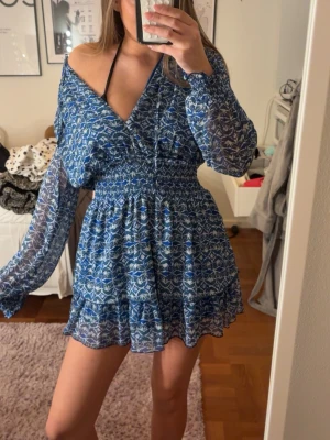 Blå mönstrad kortklänning med v-ringning - Säljer en blå kortklänning med vitt mönster och v-ringning. Klänningen har långa, transparenta ärmar och smockad midja som ger en snygg passform. Kjolen är volangig och tyget känns lätt och luftigt, perfekt för varmare dagar.