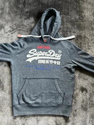Grå Superdry hoodie  - Snygg grå hoodie från Superdry med stor vit och röd logga samt japansk text på bröstet. Klassisk känguruficka framtill och justerbar huva med vita snören. Tillverkad i mjukt bomullsmaterial för skön känsla. Perfekt för en avslappnad streetwear-look.