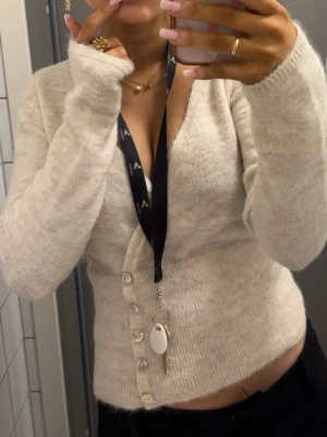 Beige kofta med sned knäppning - Trendig beige kofta med sned knäppning och pärlemorknappar. Djup V-ringning och lång ärm i mjukt, stickat material. Perfekt att styla med både jeans och kjol för en clean look.