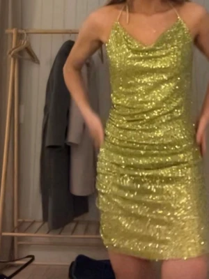Glittrig gul kort klänning - Säljer en glittrig grön kort klänning med smala band och draperad urringning. Klänningen har en figurnära passform och är täckt av paljetter som ger ett riktigt statement-utseende. Perfekt för dig som vill sticka ut och glänsa.