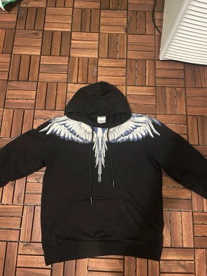 Svart hoodie från Marcelo Burlon - Säljer en svart hoodie från Marcelo Burlon med ikoniskt vingeprint i vitt och grått över axlar och bröst. Hoodien har stor huva, känguruficka. Perfekt för dig som vill sticka ut med en unik och trendig design. Mycket fint skick. Hör av dig vid frågor/ Max