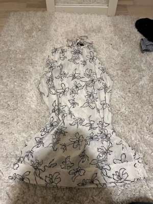 Vit halterneck med blommönster - Säljer en vit halterneck-blus med svart blommönster. Tröjan har knytning i nacken och ärmlös design, vilket ger en luftig och somrig känsla. Materialet är lätt och luftigt, perfekt för varma dagar.