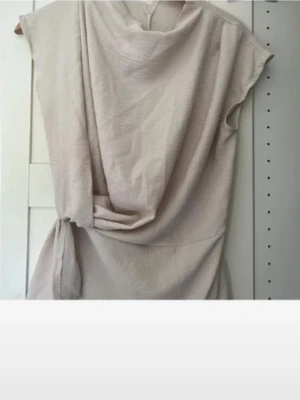 Topp - Säljer en ljusbeige omlottopp med korta ärmar och snygg drapering framtill. Toppen har ett knytband i midjan som ger en fin siluett och är tillverkad i ett mjukt material som känns skönt mot huden.