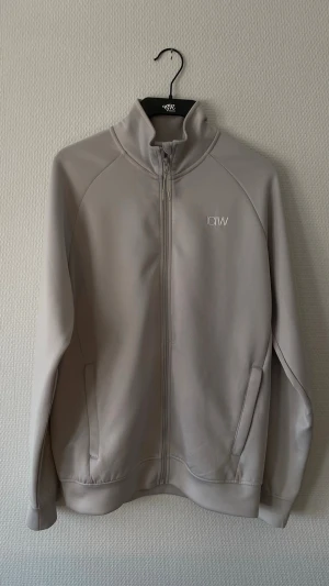 Grå ziptröja ICANIWILL - Snygg beige ziptröja från ICANIWILL med hög krage och diskret logga på bröstet. Tillverkad i ett mjukt och stretchigt material, perfekt för träning. Två fickor framtill och hel dragkedja. Enkel och stilren design.