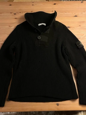 Svart stickad tröja från Stone Island - Säljer en svart ribbstickad tröja från Stone Island med hög krage och knappdetaljer framtill. Tröjan har lång ärm och den klassiska Stone Island-loggan på vänster ärm. Perfekt för dig som gillar streetwear och snygga detaljer.